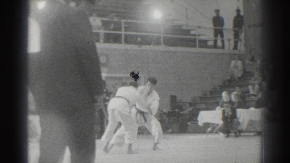 The karate match continues on the raised platform, with competitors bent forward and officials or spectators visible around the arena.