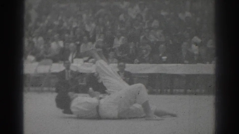 Black-and-white 1966 San Gabriel home-movie frame showing two martial artists on a mat, with one competitor down and a blurred audience behind.