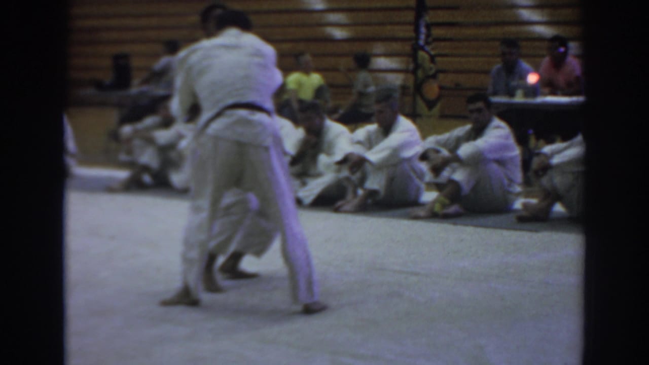 Two martial artists grapple on a gym mat while seated competitors watch from the sidelines.