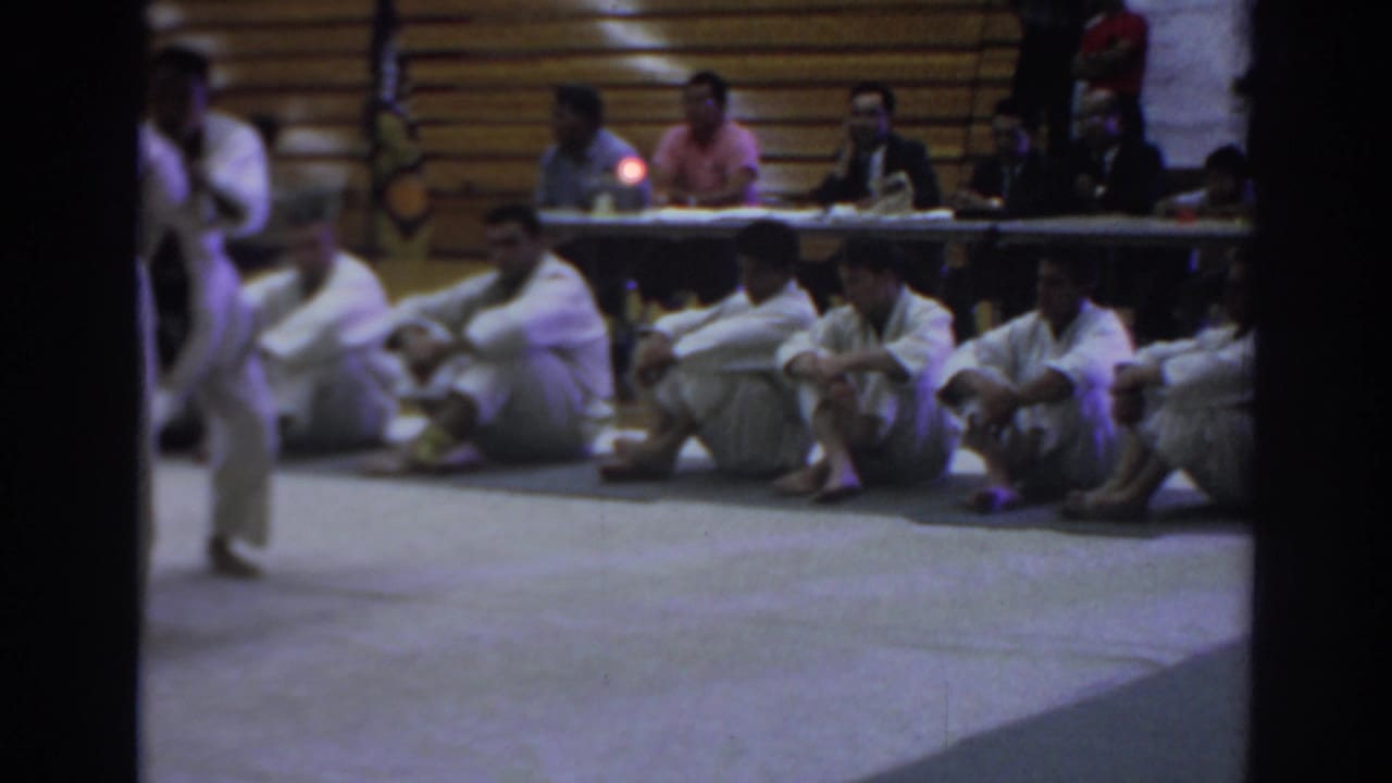 Uniformed competitors sit beside a gym mat as officials watch from a table near the bleachers.