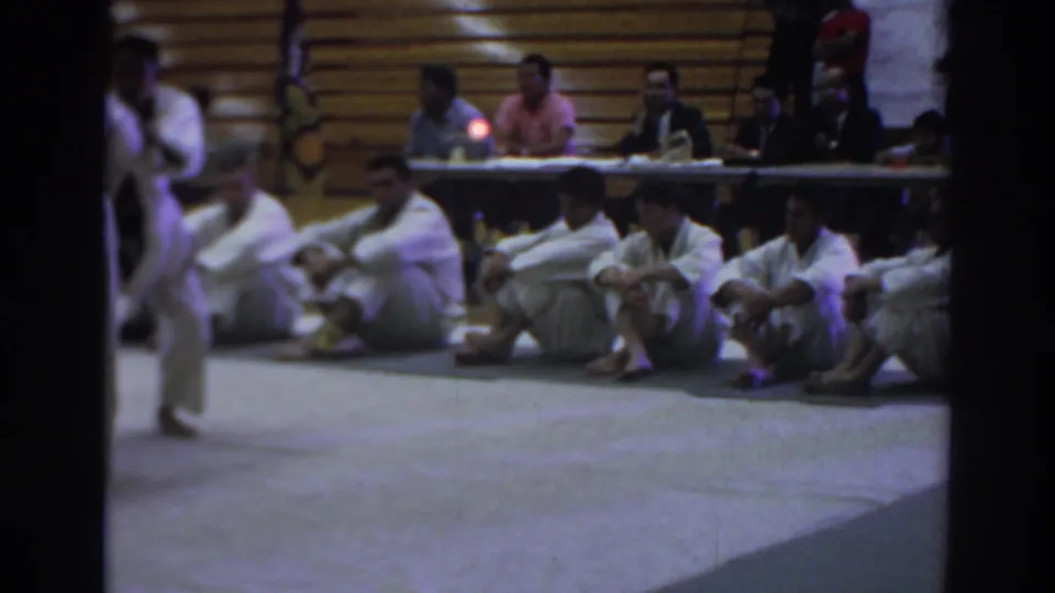 Temple City gym scene with seated martial arts competitors, judges at a table, and a match continuing partly out of frame in 1966.