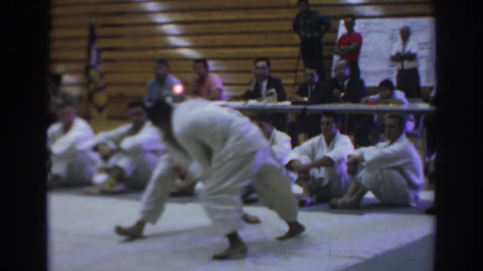 A 1966 Temple City judo competitor bends toward the mat during a match, with seated participants, officials, and bleachers behind him.