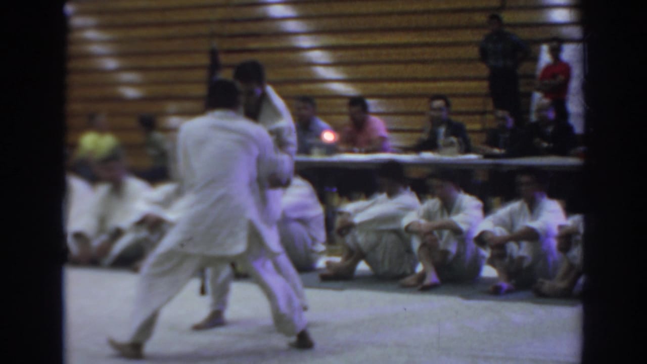 Two karate competitors spar on a gym floor while spectators sit along a wooden wall.