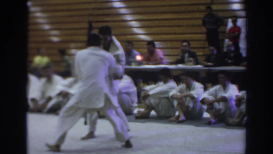 1966 Temple City home-movie frame showing two karate competitors in white gi sparring before seated spectators in a gymnasium.