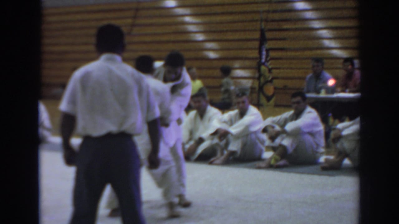 A referee observes two karate competitors in white gi during a 1966 Temple City gymnasium sparring match.