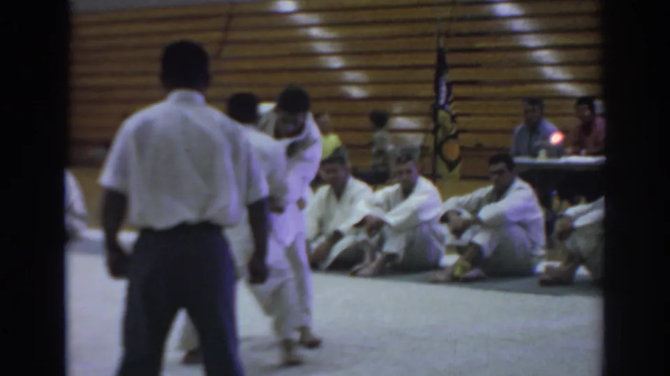 A referee observes two karate competitors in white gi during a 1966 Temple City gymnasium sparring match.