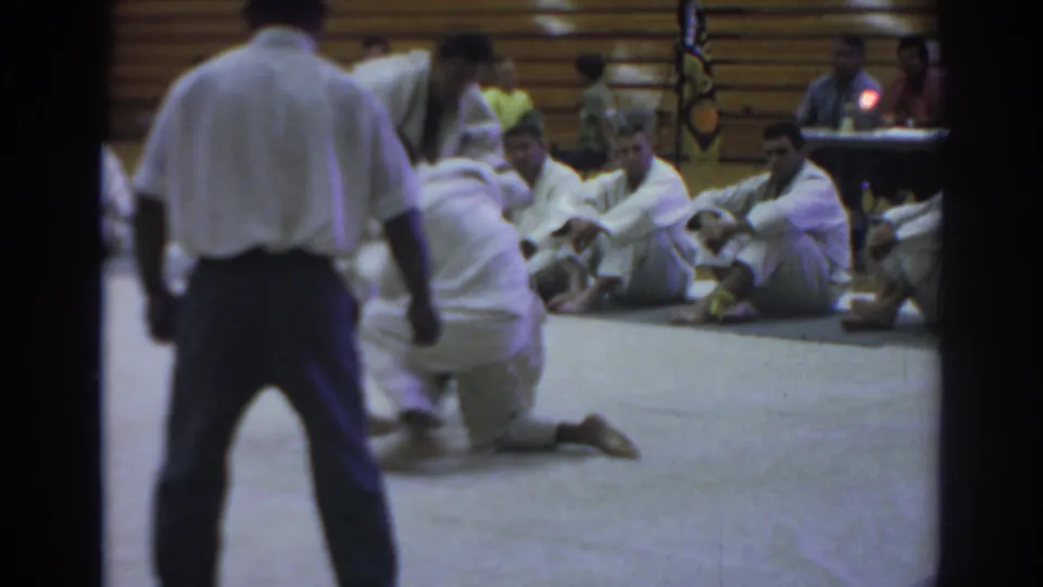 Temple City gymnasium frame from 1966 showing a karate competitor on one knee during a sparring exchange with the referee in view.
