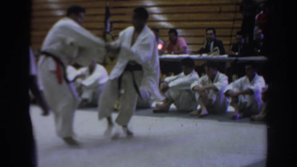A 1966 Temple City home-movie frame shows two karate competitors in white gi facing off beside seated spectators and a judges table.