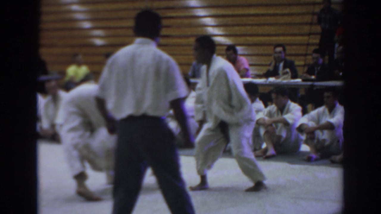 Two judo athletes grapple on a gym mat while a referee stands nearby.