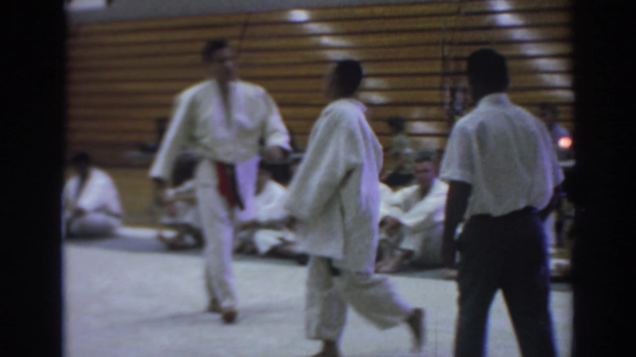 Temple City 1966 frame of two judo competitors in white gi moving on the mat with a referee and seated participants along the gym wall.
