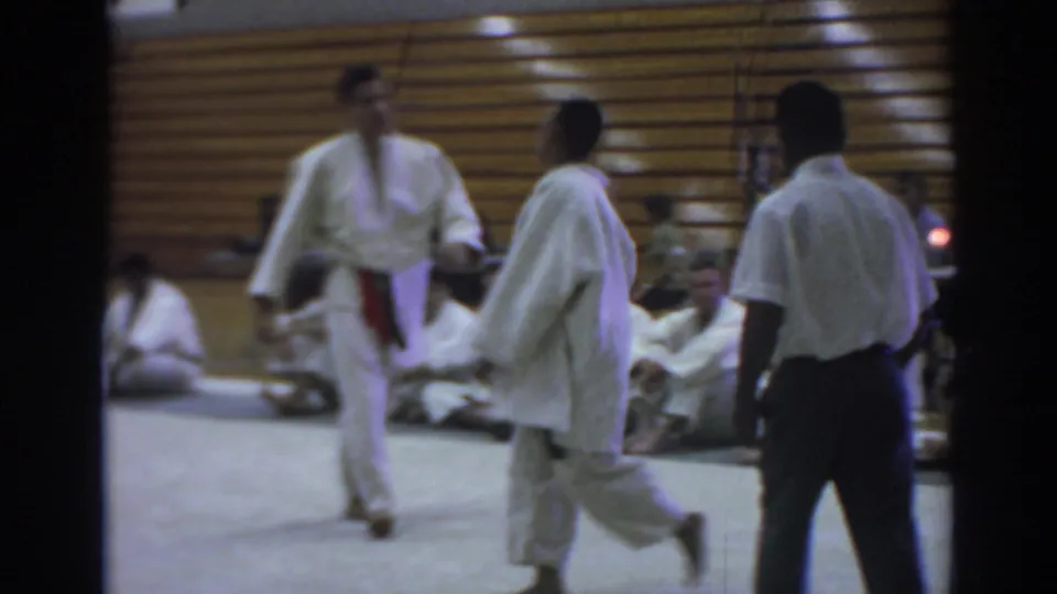 Temple City 1966 frame of two judo competitors in white gi moving on the mat with a referee and seated participants along the gym wall.