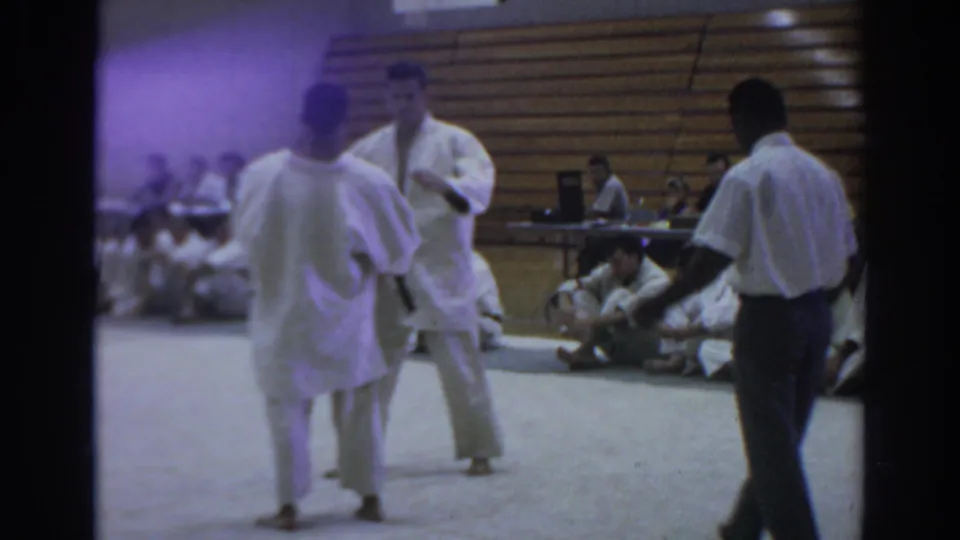 Temple City 1966 judo match frame with two athletes facing on the mat, a referee nearby, and competitors seated against wooden bleachers.