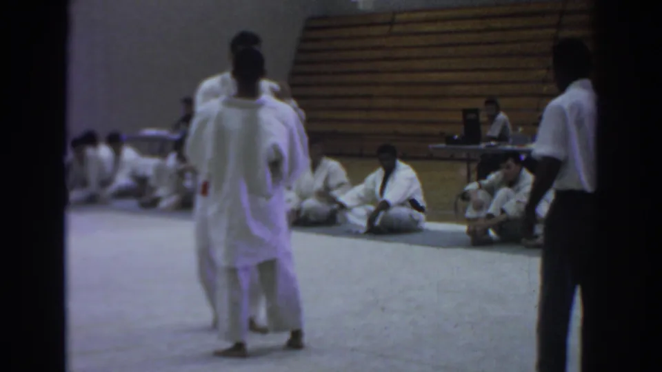 Temple City 1966 frame showing judo athletes in white gi on a gym mat, with a referee at right and competitors seated along the wall.