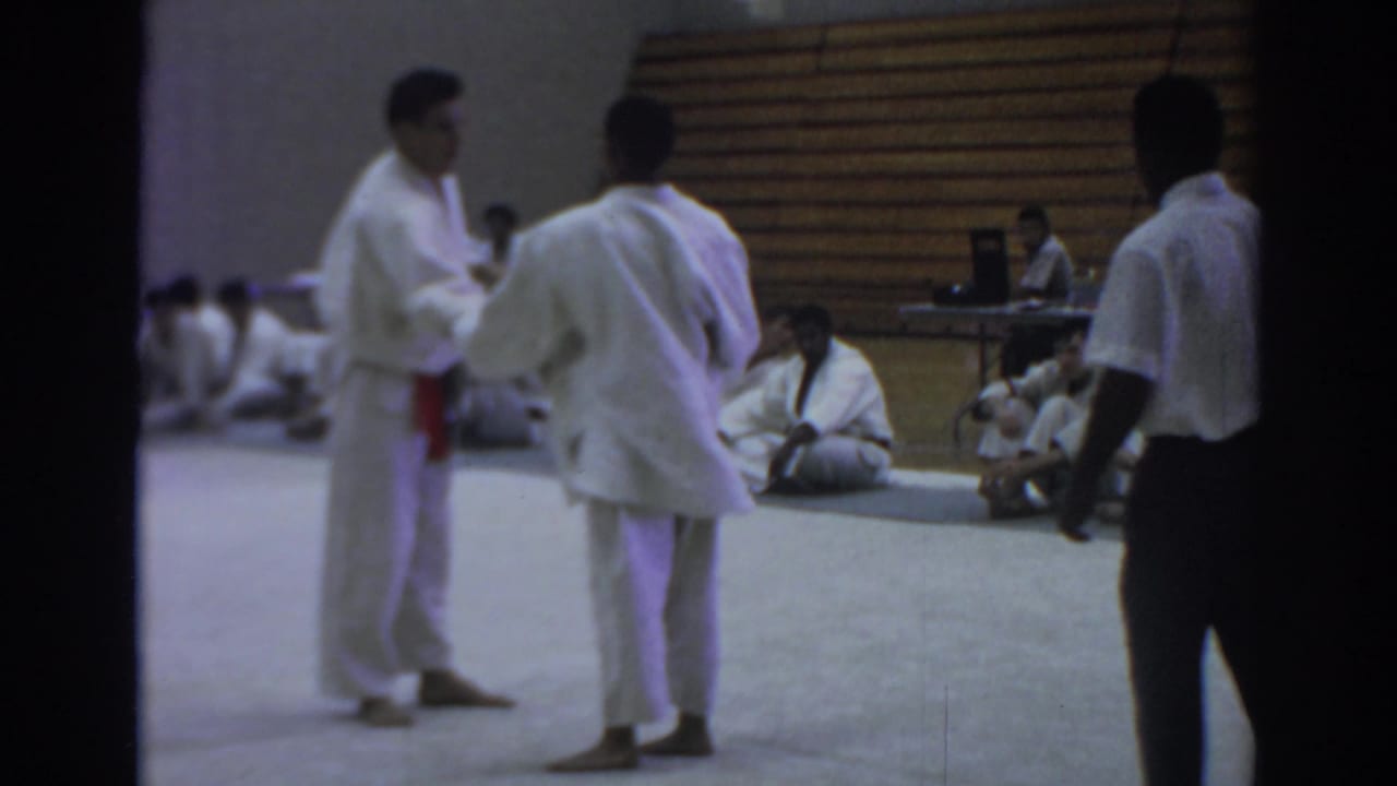 In 1966 Temple City home-movie footage, two judo participants in white gi stand on a mat as seated observers watch from the gym floor.