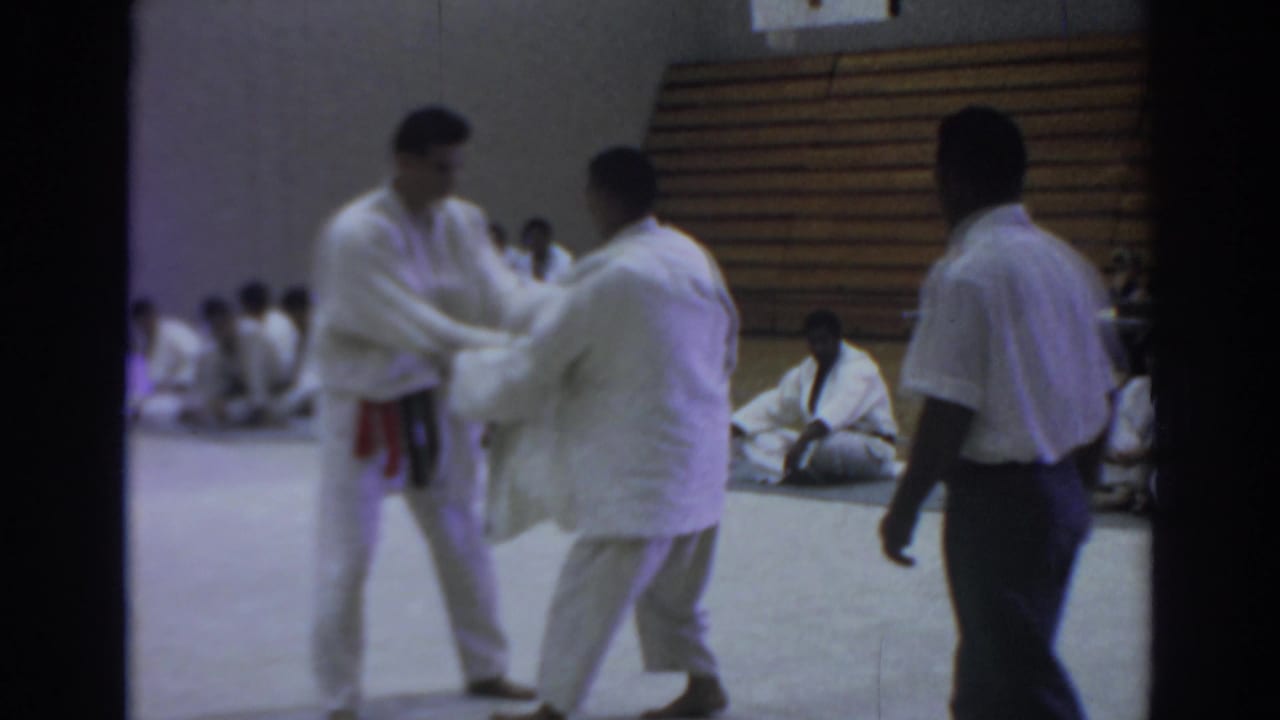 Two judo participants grip each other's sleeves as a man stands near the mat.
