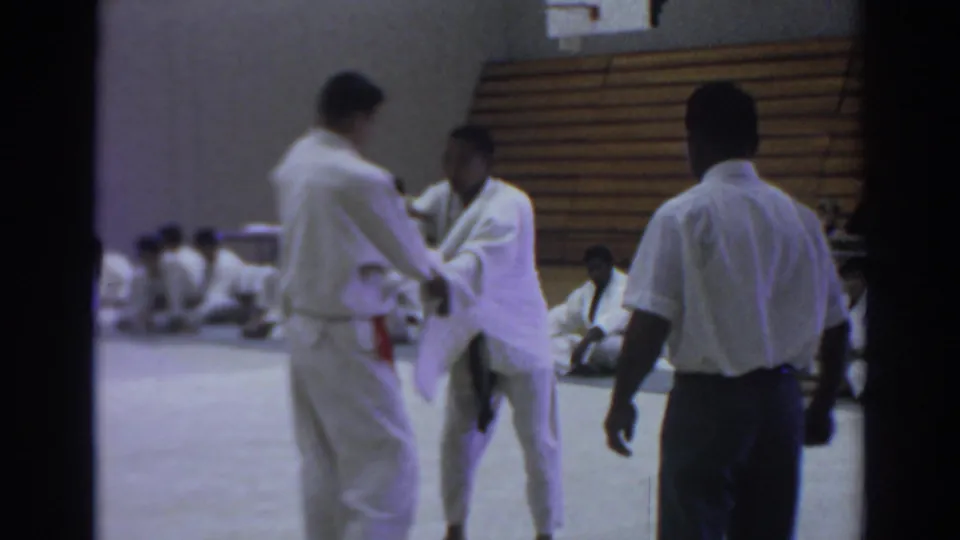 During the short 1966 judo sequence, one participant steps inward while holding the other man's sleeve or arm in the gymnasium.