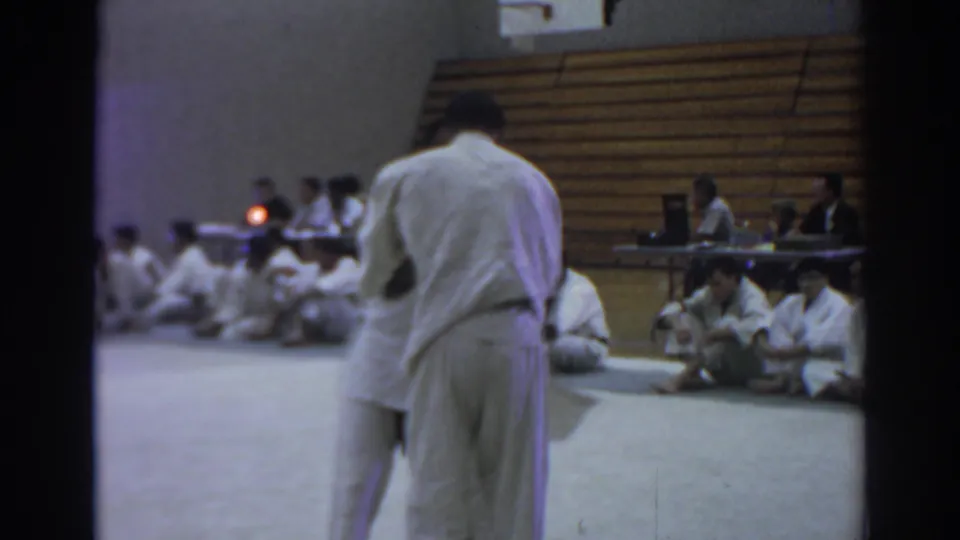 The pair rotates in a judo grapple on the mat, with seated participants, bleachers, and a side table visible in the Temple City gym.