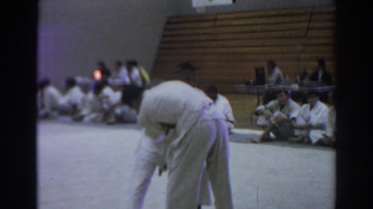 Two judo competitors in white uniforms grapple on a gym mat while spectators sit near bleachers.