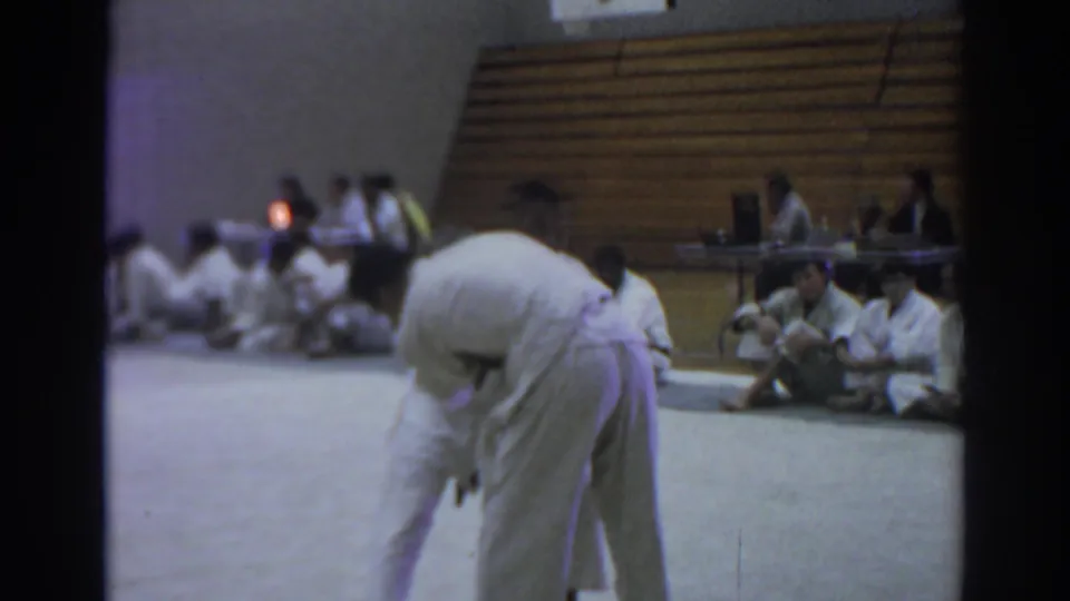 1966 Temple City gym scene with two judo competitors in white gi uniforms grappling on a light mat as seated spectators watch.