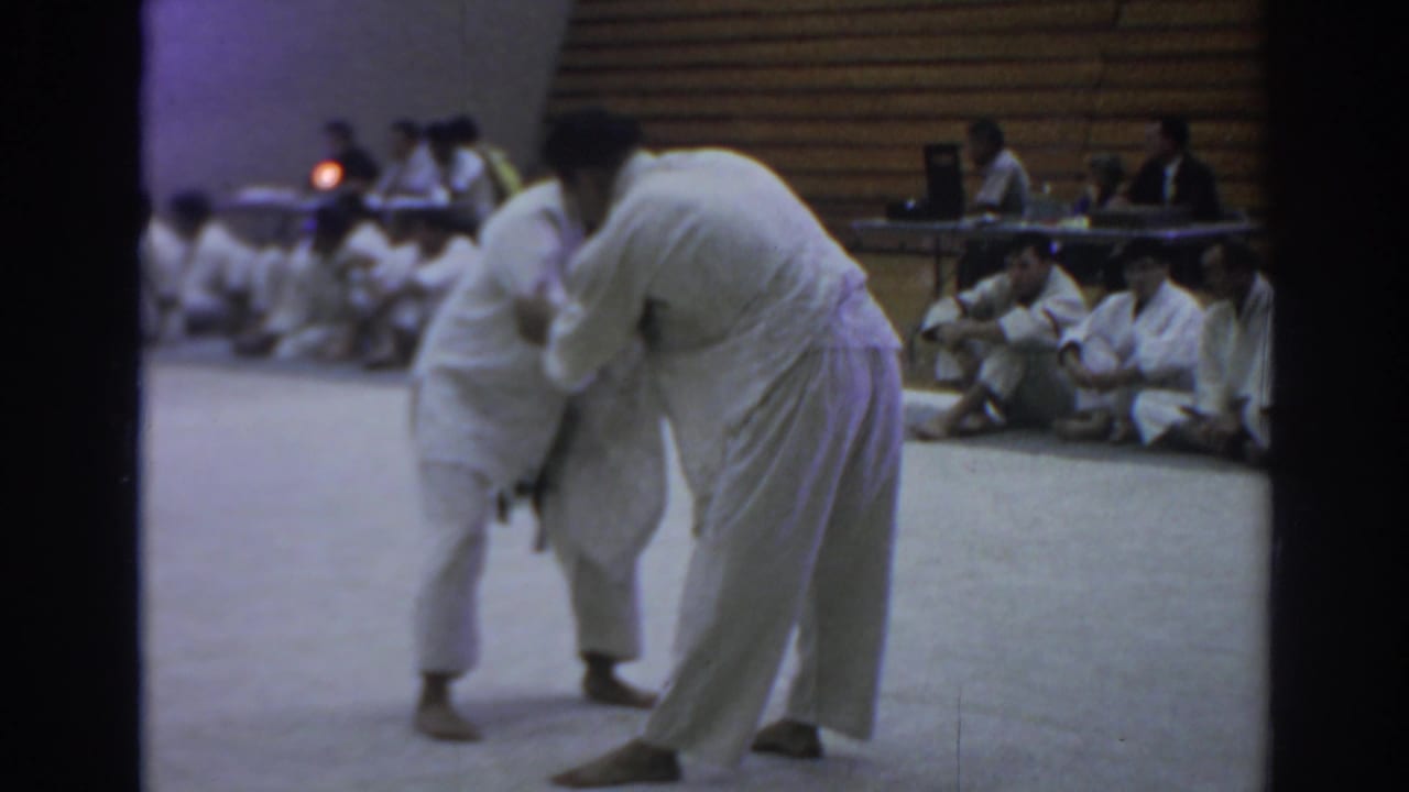 Two judo athletes grapple upright on a light-colored mat in a 1966 Temple City gym, with seated participants and officials behind them.