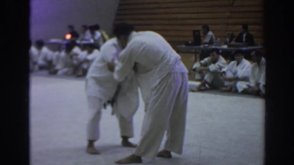 Two judo athletes grapple upright on a light-colored mat in a 1966 Temple City gym, with seated participants and officials behind them.
