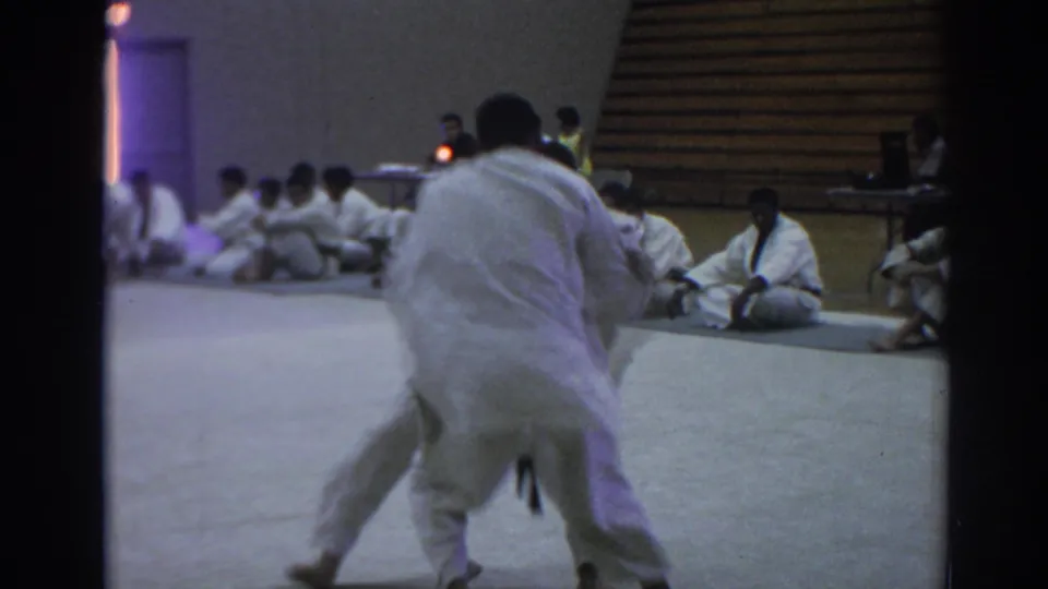 A competitor in a white gi moves across the mat during a Temple City judo match, with other uniformed participants seated along the wall.