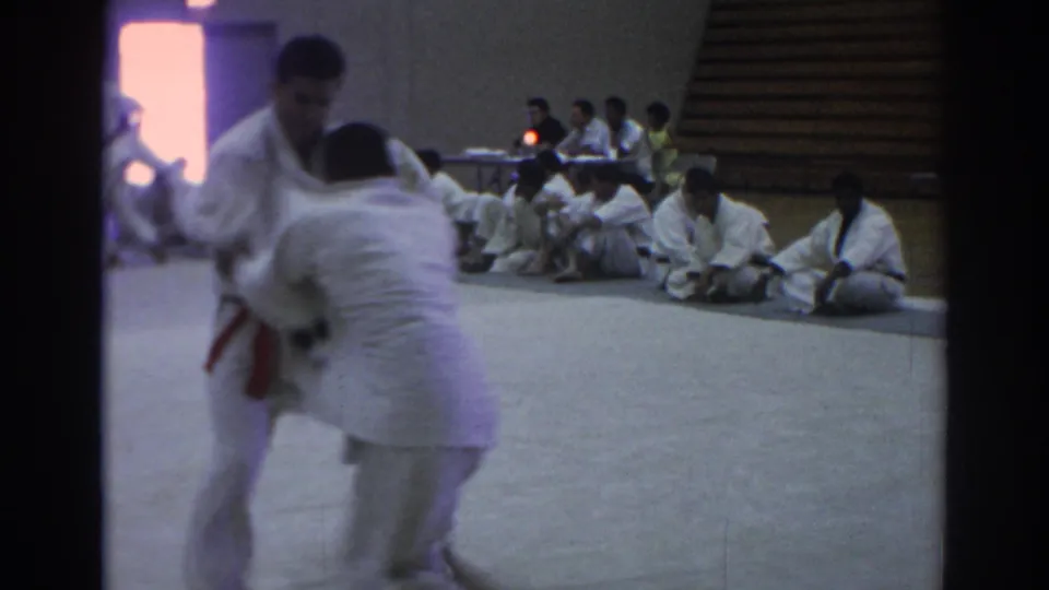 Two judo competitors in white uniforms grapple on a gym mat in Temple City, California, with seated spectators and officials visible behind them.