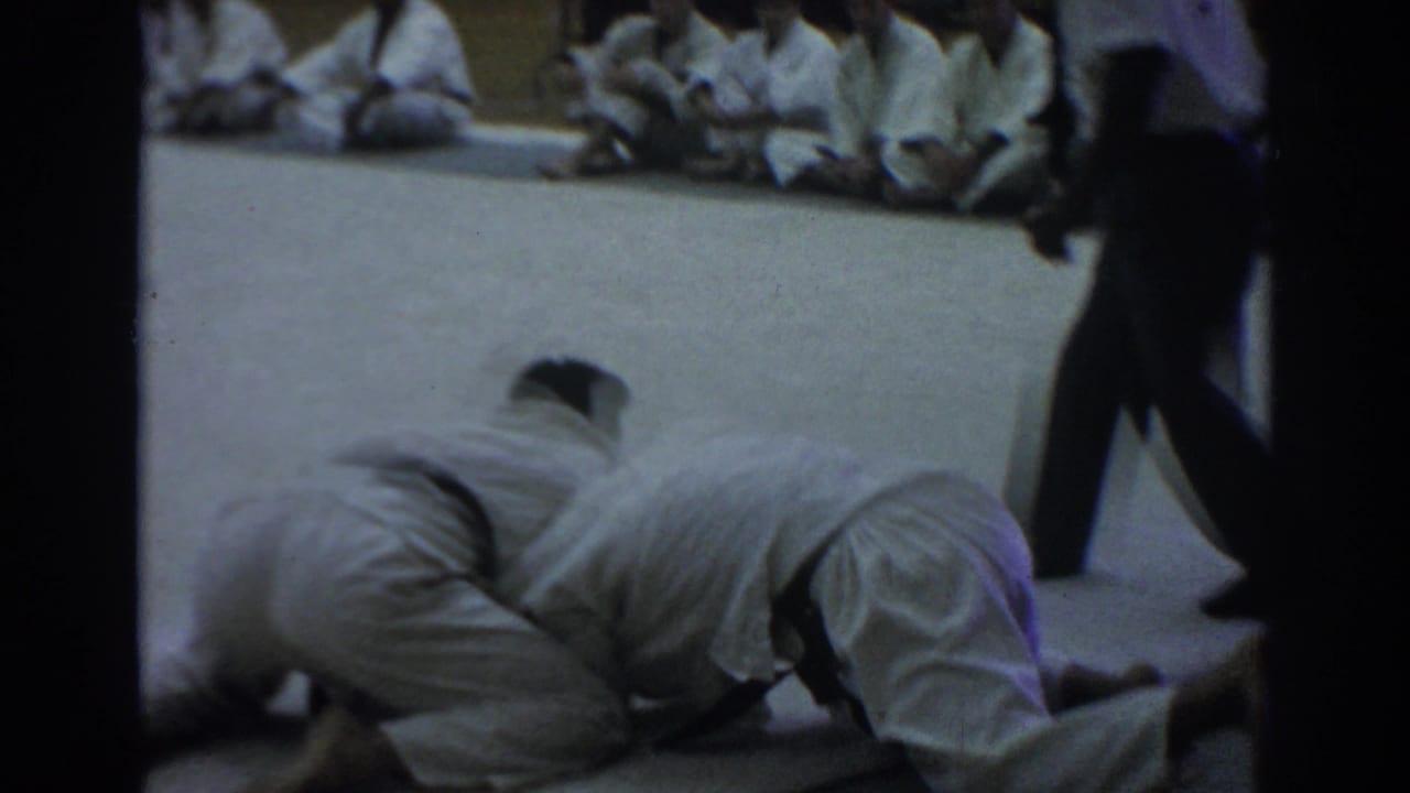 Two martial artists grapple low on the mat as seated participants watch nearby.