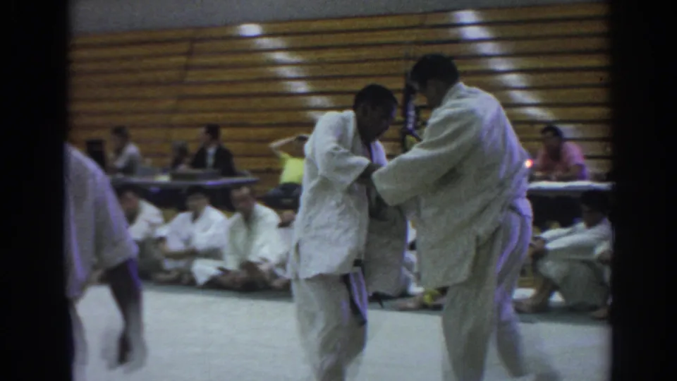 In the 1966 Temple City gym scene, two gi-clad competitors grip each other while seated participants and officials watch from behind.