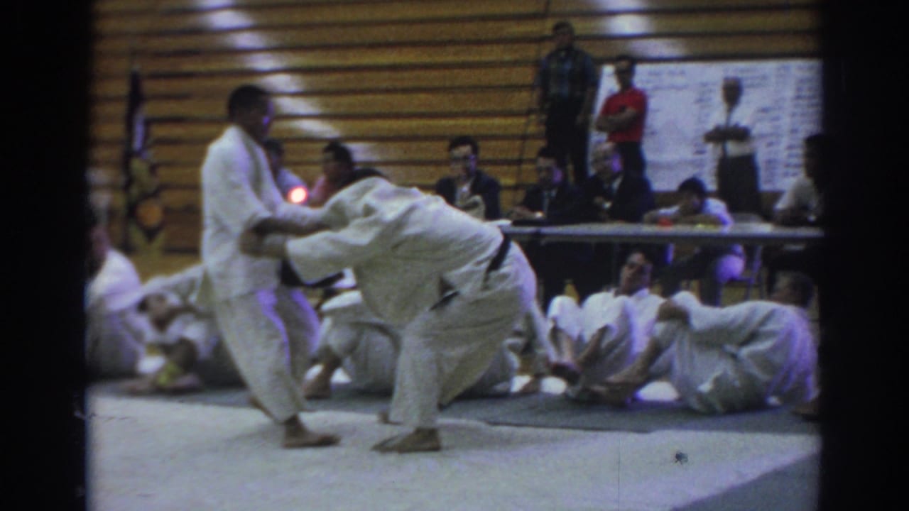 Two judo competitors grapple on a gym mat while seated participants watch near bleachers.