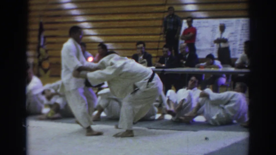 1966 Temple City home-movie frame showing two judo competitors in white gi during a match, with seated participants and judges behind them.