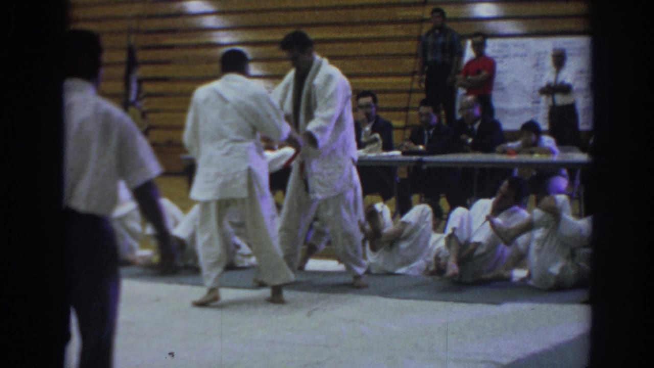 Judo competitors stand on the mat as others sit along the wall near a judges' table.