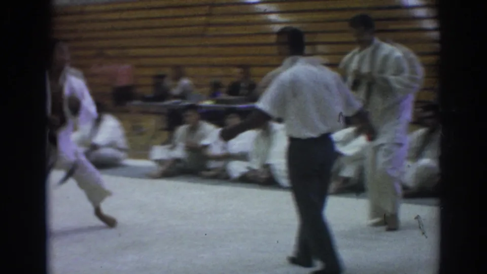 1966 indoor judo footage shows a referee crossing the mat between competitors, with seated martial arts participants against the bleachers.