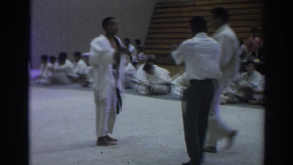 Temple City 1966 judo competition frame with a competitor in a white gi facing the referee and other participants seated along the gym wall.
