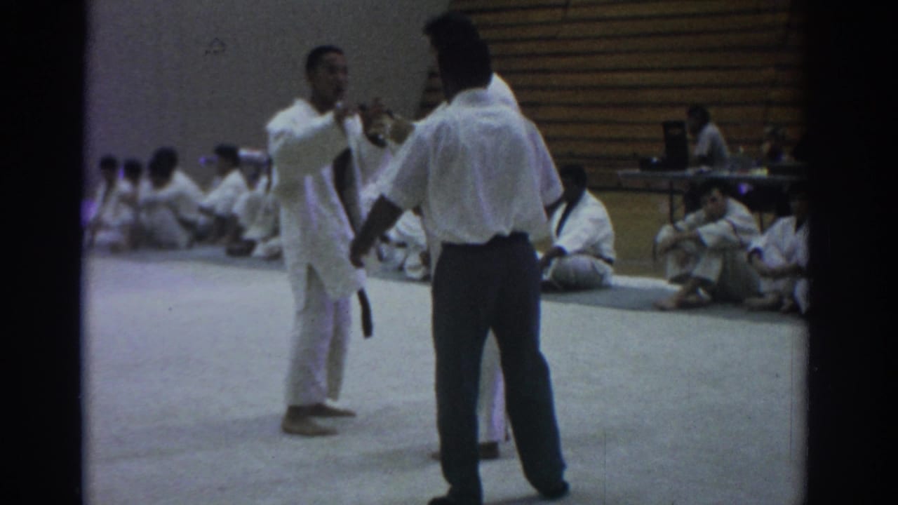 Judo athlete and referee stand on a gym mat while seated participants watch from the sideline.