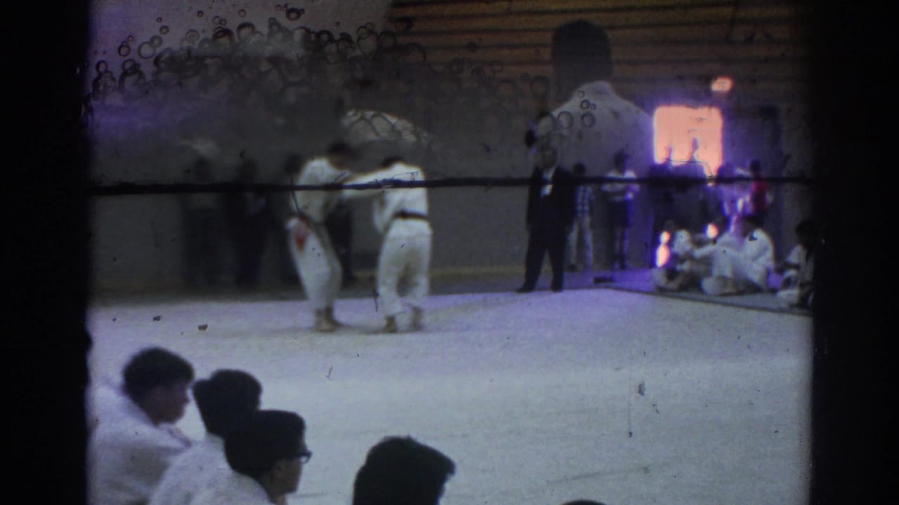 Two judo athletes grapple on a mat as spectators and participants sit along the gym wall.