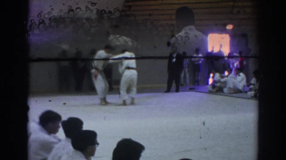 Two athletes in white gi grapple during a 1966 Temple City judo match, with seated observers and a bright doorway behind them.