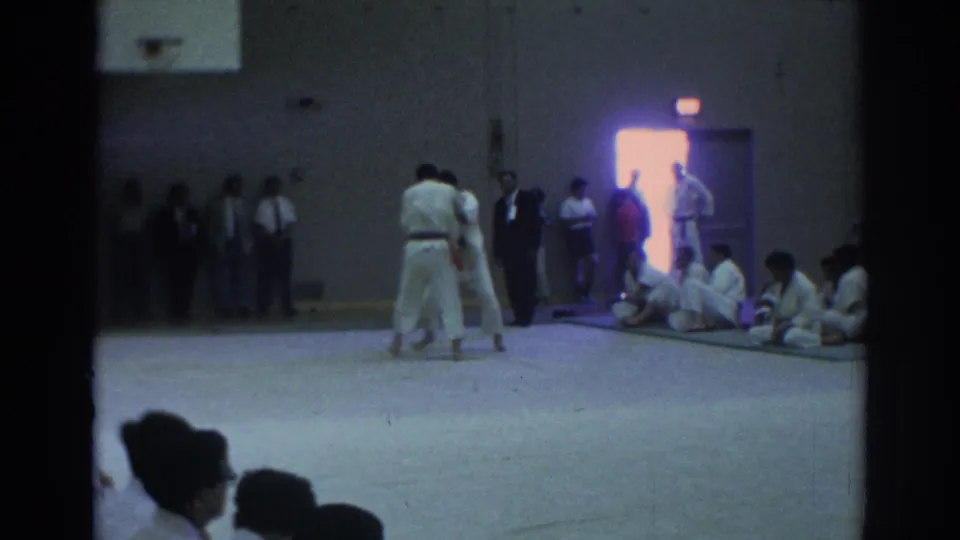 Two white-gi judo athletes stand in a Temple City gym while seated participants, officials, and spectators watch from the sidelines.
