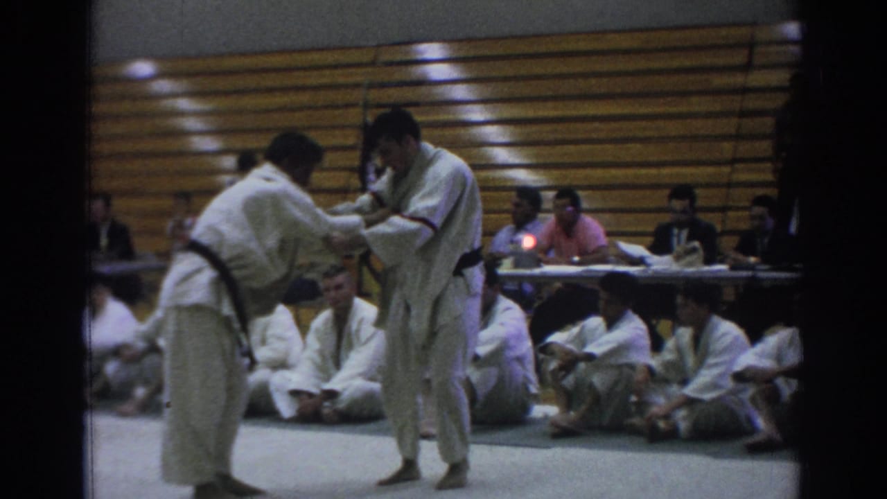 In Temple City, California, 1966, two judokas in white gi grapple on a gymnasium mat as spectators sit near wooden bleachers.