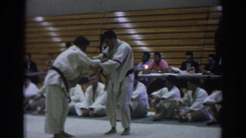 In Temple City, California, 1966, two judokas in white gi grapple on a gymnasium mat as spectators sit near wooden bleachers.