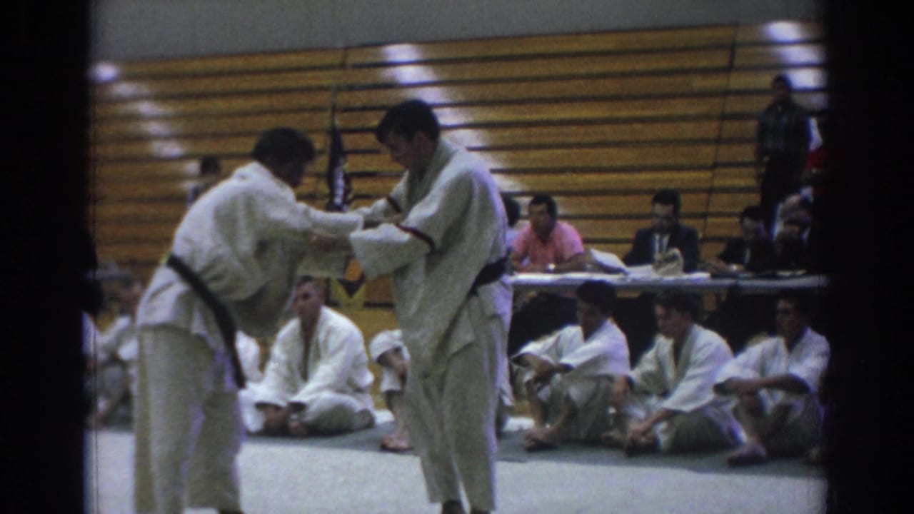 Two judo competitors hold sleeve and lapel grips in front of bleachers.