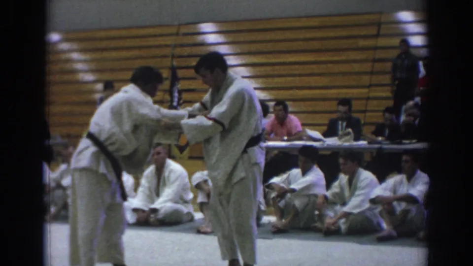 Two judo competitors in white gi hold grips during a 1966 Temple City gymnasium match, with seated observers and bleachers behind them.