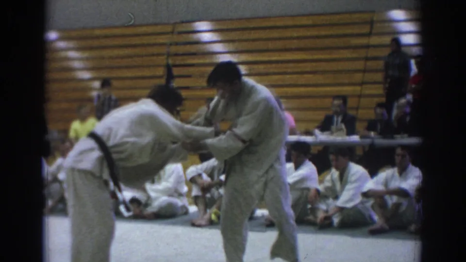 A judoka bends low during a Temple City judo bout as the opponent controls the grip, with seated participants watching from the mat edge.