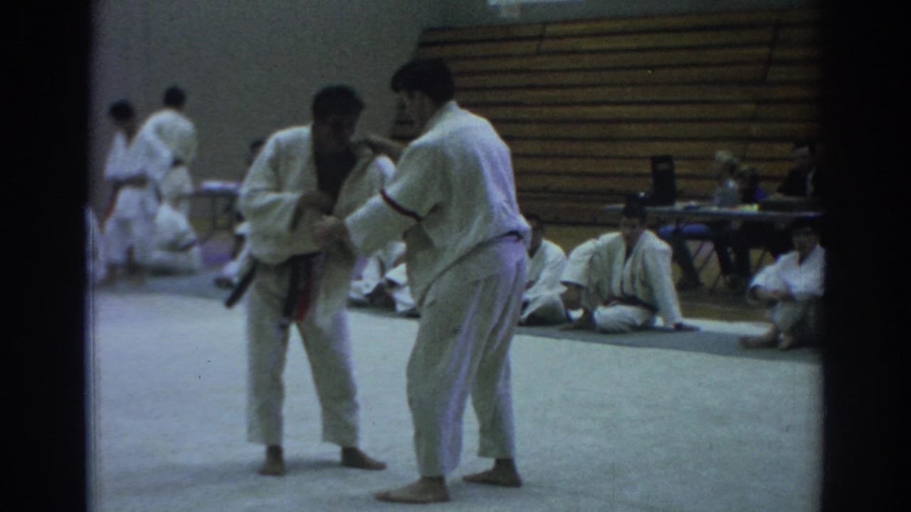 Two martial artists in white gi practice on a gym mat as others sit nearby.