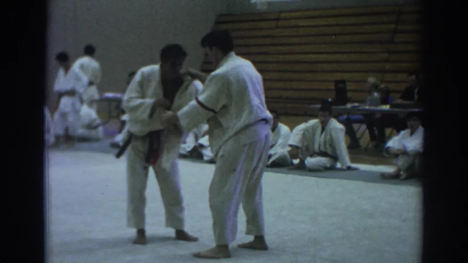 1966 Temple City gym scene with two martial artists in white gi demonstrating close-quarters technique before seated students.