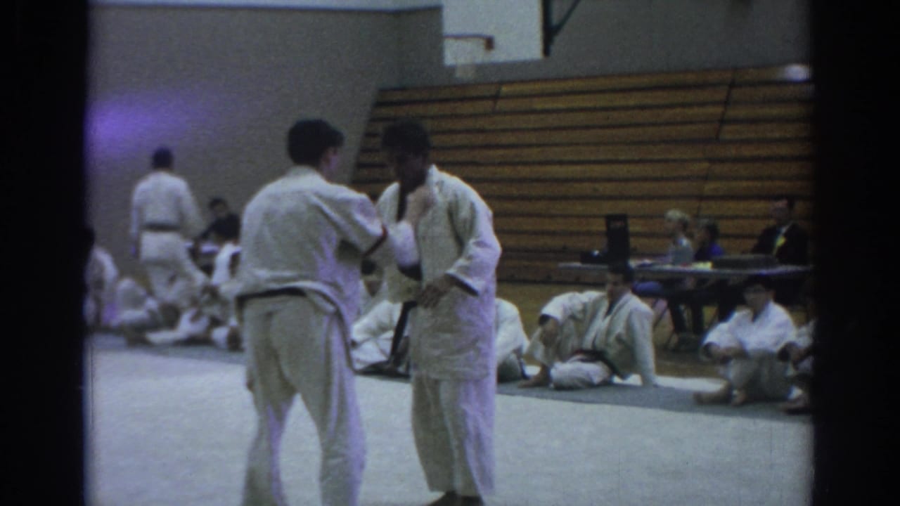 At the Art Macias Academy demonstration, two practitioners stand in close contact on a gym mat while students watch from the floor.