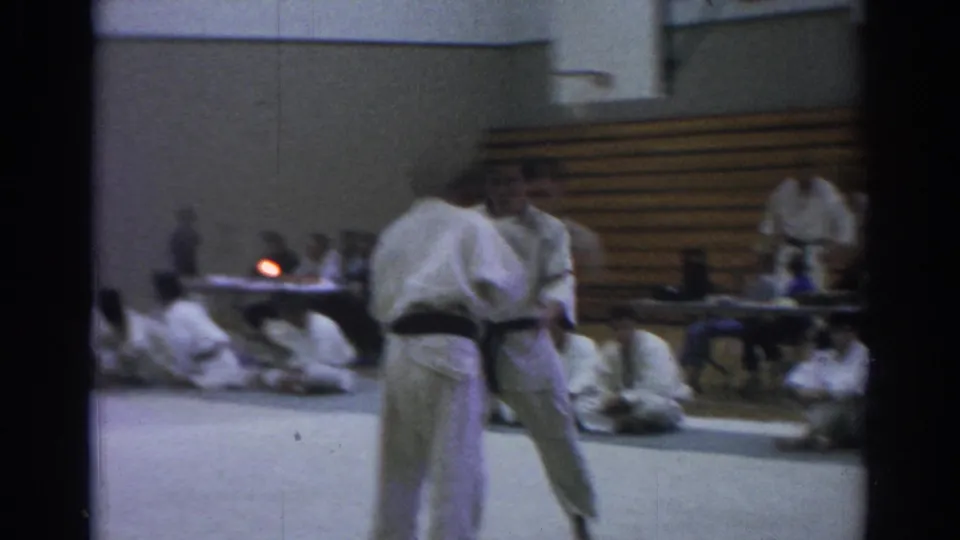 Soft 8mm frame of a Temple City martial arts demonstration with two gi-clad figures moving before seated students and bleachers.