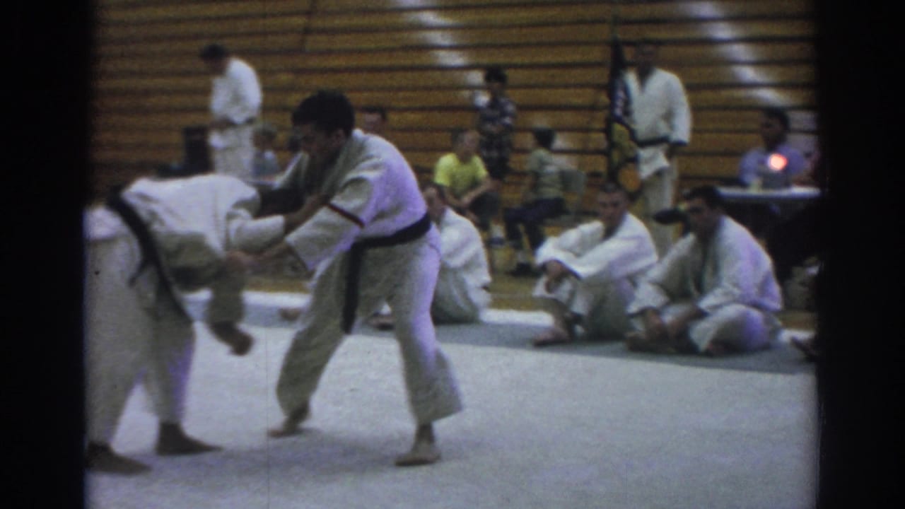 Two martial artists in white gi grapple on a mat with seated spectators behind them.