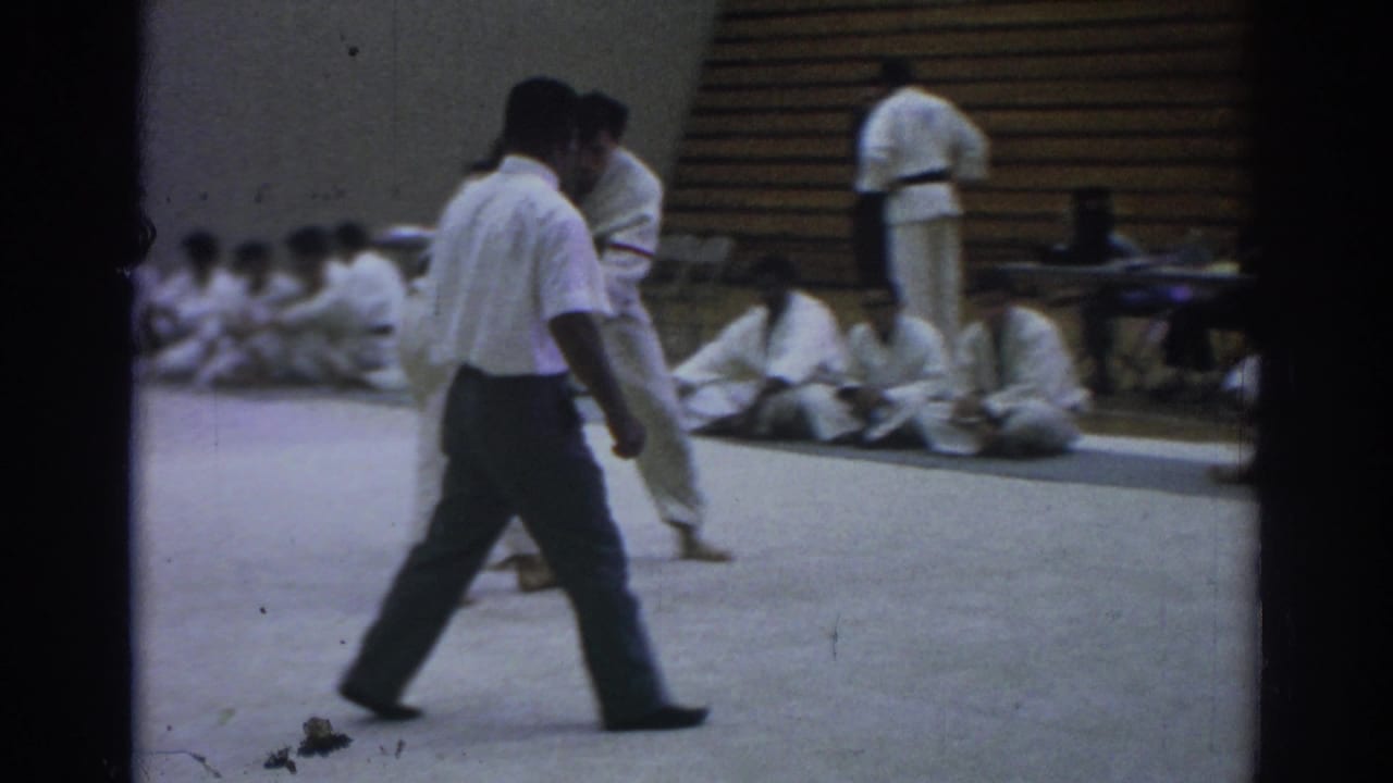 Temple City 1966 8mm frame with a referee crossing the mat during a martial arts match, with gi-clad participants seated near wooden bleachers.