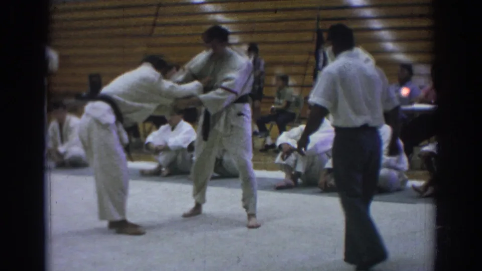 Temple City 1966 8mm frame of a jujitsu match with one competitor leaning forward into a standing opponent while the referee observes.